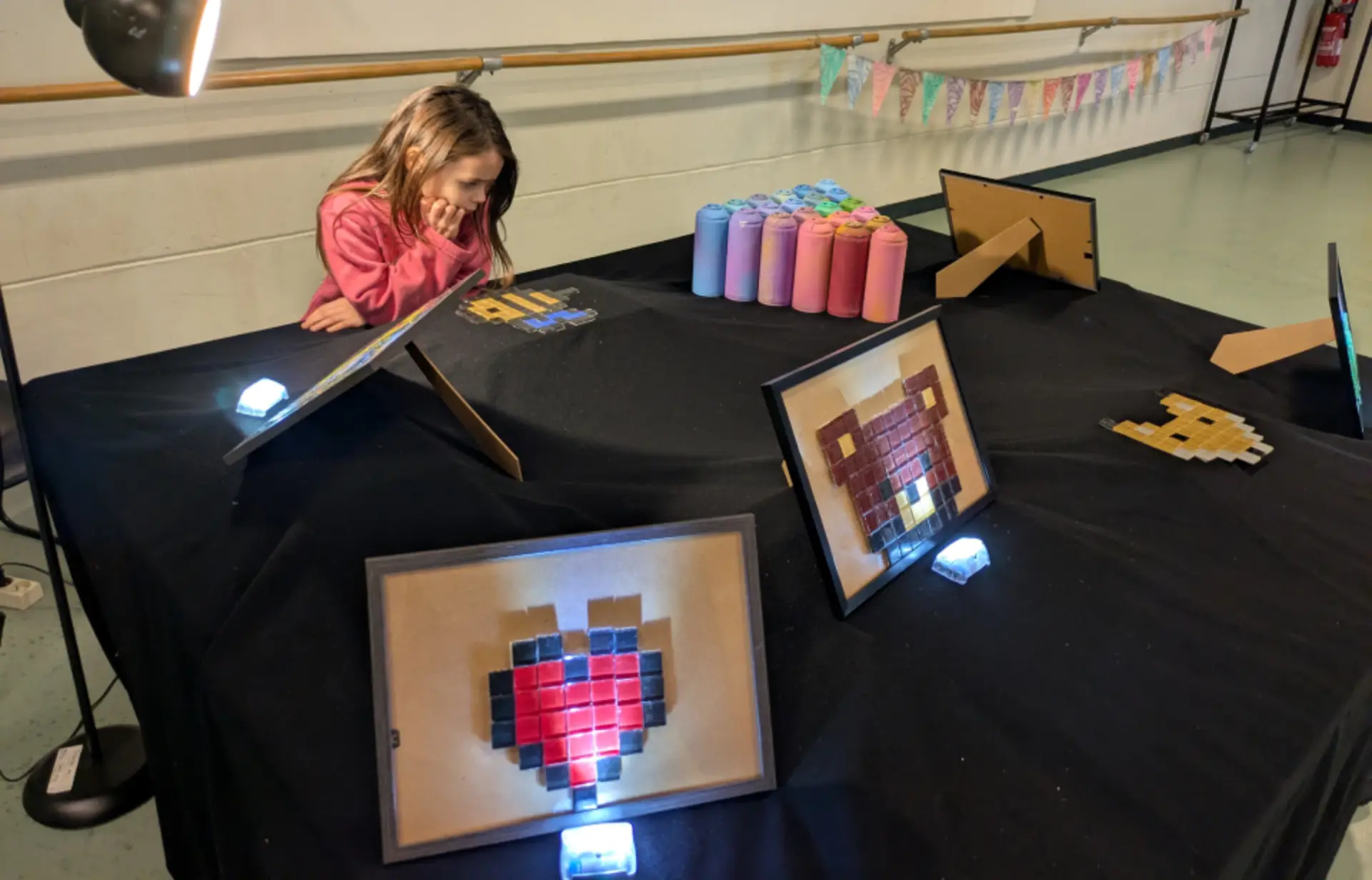 Table couverte d'un tissu noir, exposant des pièces de pixel art encadrées. Trois pièces sont visibles : un cœur rouge, un visage d'ours et un design jaune et bleu. Plusieurs bombes de peinture de différentes couleurs sont disposées sur la table. Un enfant portant un pull rose se tient derrière la table, regardant le pixel art. La table est éclairée par une lampe sur le côté gauche. En arrière-plan, il y a une bannière colorée et un porte-manteau.